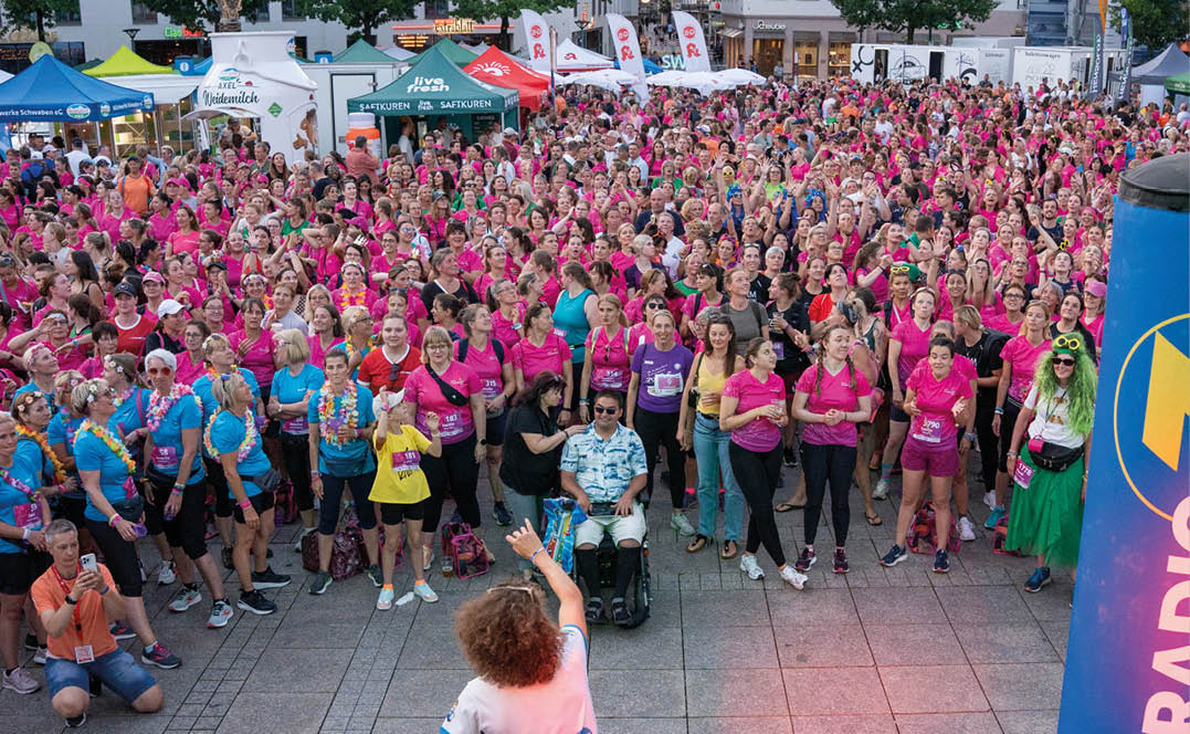 A large crowd of people wearing colorful shirts gathers outdoors while a person with curly hair in a white outfit speaks to them using a microphone. Beschreibung von KI generiert