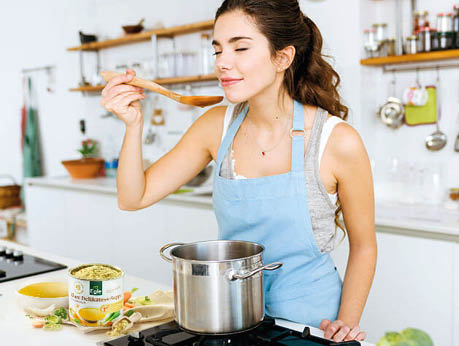 A person wearing a blue apron is tasting food from a wooden spoon while standing in a bright kitchen with a pot on the stove and a container of seasoning nearby. Beschreibung von KI generiert