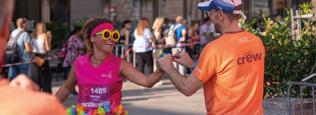 A person wearing a bright pink shirt and colorful tutu is holding hands with another person in an orange shirt and cap during an outdoor event with a crowd in the background. Beschreibung von KI generiert
