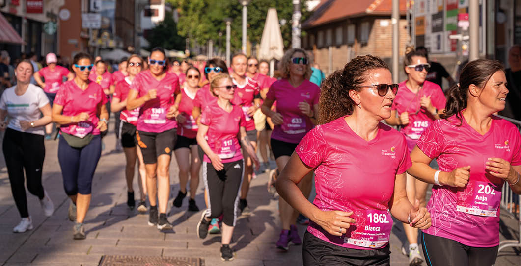 A group of people wearing matching pink shirts and sunglasses are running together on a sunny street in an organized event. Beschreibung von KI generiert
