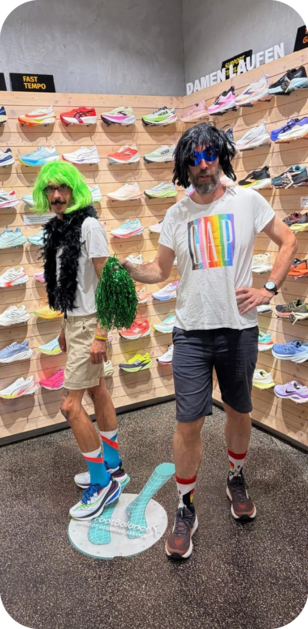 Two people with colorful wigs and casual clothing pose playfully in a shoe store with walls covered in various athletic shoes. Beschreibung von KI generiert