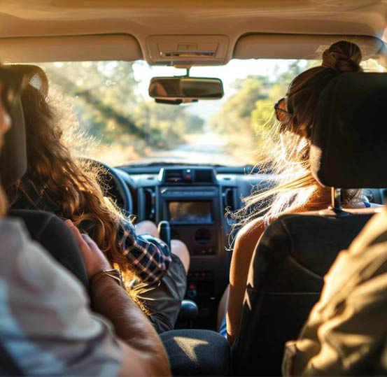 Three people are sitting inside a car with one person driving on a sunlit road surrounded by greenery. Beschreibung von KI generiert