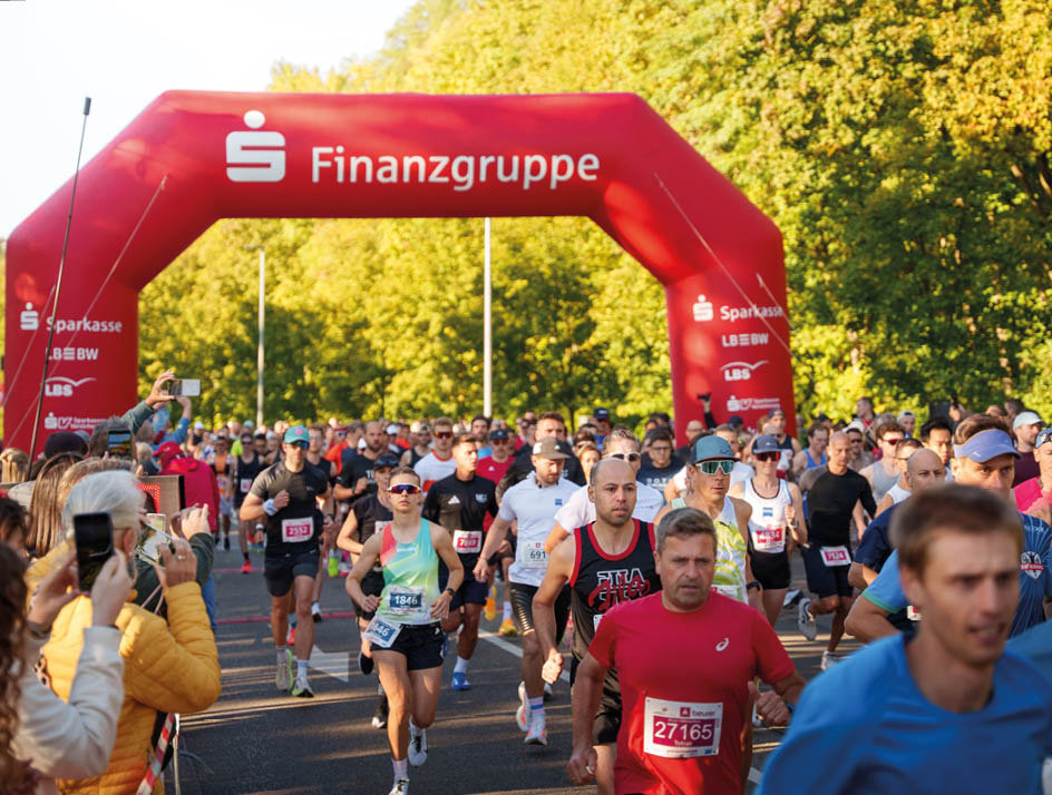 A large group of people wearing athletic clothing are running on a road during a race event with spectators on the sides. Beschreibung von KI generiert