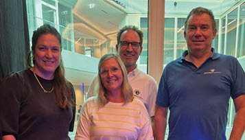 Four adults smiling and standing closely together indoors with large windows and modern architecture in the background. Beschreibung von KI generiert