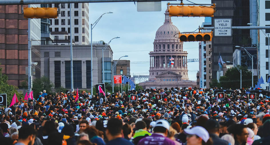 A large crowd of people gathers in a city street with the Texas State Capitol building visible in the background under a cloudy sky. Beschreibung von KI generiert