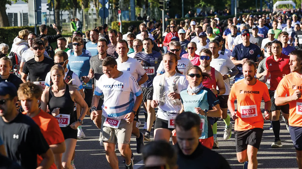 A large group of people wearing athletic clothing and numbered bibs are running together in an outdoor race on a sunny day. Beschreibung von KI generiert