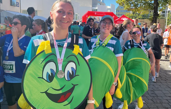 Three adults wearing green costumes with smiling faces and yellow gloves are standing in a row outdoors at a crowded event, with one person in front wearing a medal. Beschreibung von KI generiert