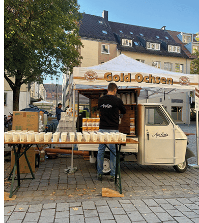 An outdoor market stall with a white canopy labeled Gold Ochsen serves drinks from a small vintage vehicle while a person in a black shirt arranges cups on a table in front. Beschreibung von KI generiert