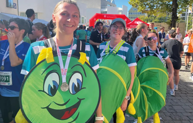 Three adults wearing green costumes with smiling faces and yellow gloves are standing in a row outdoors at a crowded event, with one person in front wearing a medal. Beschreibung von KI generiert