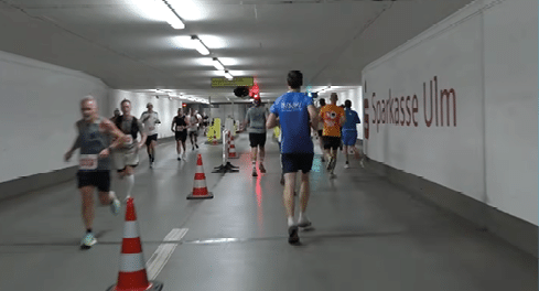 People wearing athletic clothing are running through a well-lit tunnel with traffic cones and a sign that reads “Sparkasse Ulm" on the wall. Beschreibung von KI generiert