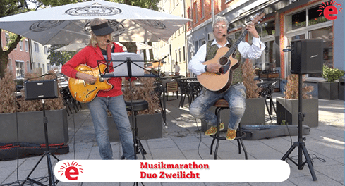 Two adults are playing guitars and singing outdoors on a city street with tables and chairs in the background during a music event. Beschreibung von KI generiert