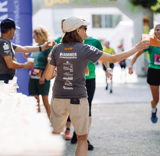 A person wearing a cap and a crew t-shirt is handing a cup of water to a runner during an outdoor race event. Beschreibung von KI generiert