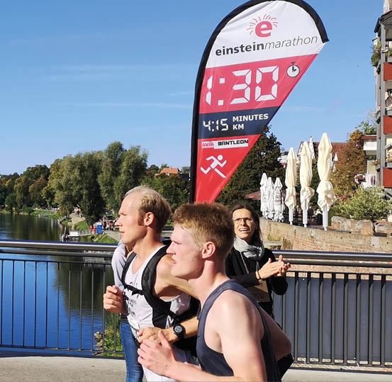 Two adults are running outdoors near a river with a large red and white marathon timer flag in the background showing elapsed time and pace. Beschreibung von KI generiert