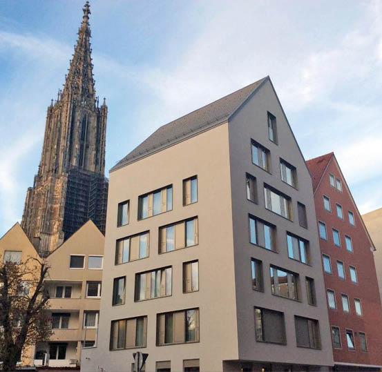 A modern multi-story building with large windows stands in front of a tall, ornate Gothic cathedral tower under a partly cloudy sky. Beschreibung von KI generiert