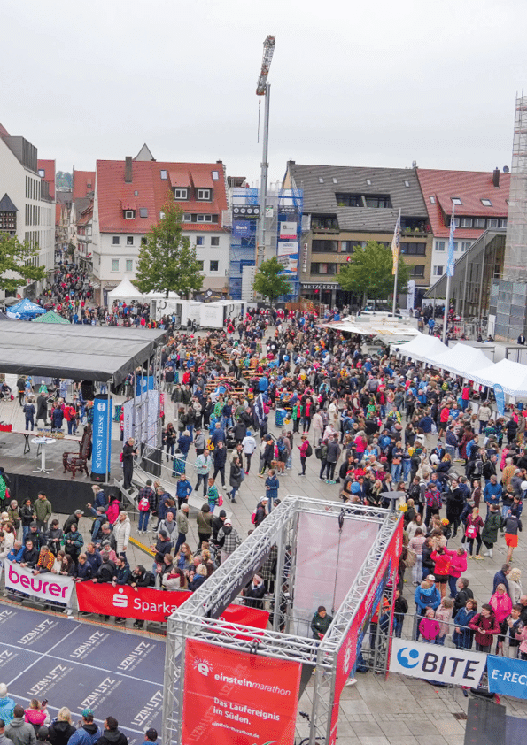 A large crowd of people gathers in an outdoor urban area with tents, banners, and a stage setup, suggesting a public event or festival. Beschreibung von KI generiert