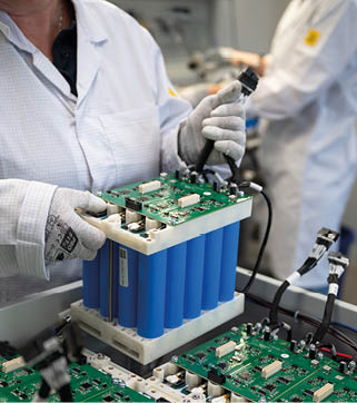 A person wearing gloves and a white lab coat is handling a large blue battery pack with circuit boards in a laboratory or workshop setting. Beschreibung von KI generiert