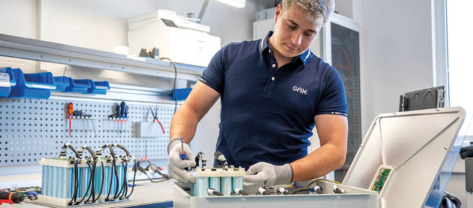 A person wearing gloves and a navy blue polo shirt is working with electronic equipment and components on a workbench in a well-lit workshop. Beschreibung von KI generiert