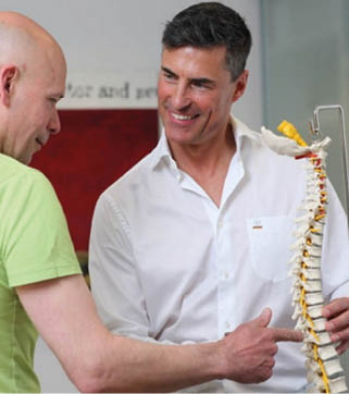 Two adults are engaged in a discussion while one points to a model of a human spine held by the other in a clinical or educational setting. Beschreibung von KI generiert
