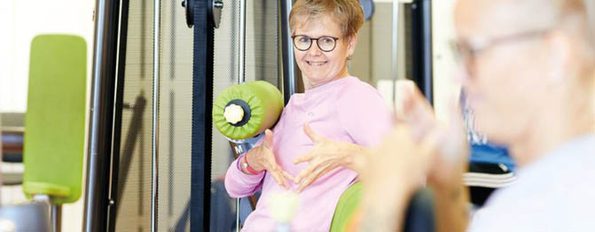 A person wearing glasses and a pink shirt is exercising on a machine while another person in the foreground is also engaged in a workout. Beschreibung von KI generiert