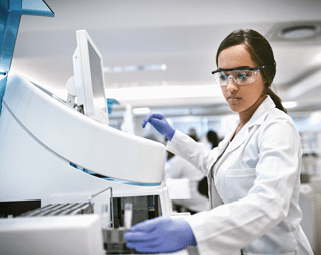 A person wearing safety goggles, a white lab coat, and purple gloves is operating laboratory equipment in a scientific setting. Beschreibung von KI generiert