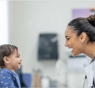 An adult with hair tied back and wearing a white coat smiles and interacts with a child wearing a blue shirt in a bright indoor setting. Beschreibung von KI generiert