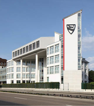 A modern white multi-story building with large windows and a distinctive vertical section featuring a black emblem and red stripe stands beside a road under a clear blue sky. Beschreibung von KI generiert