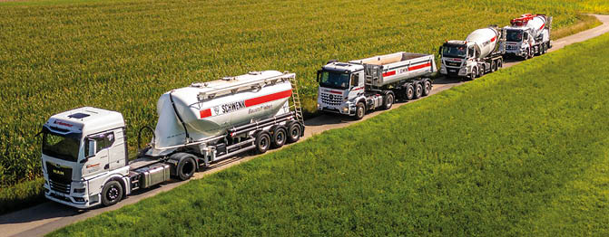 Four white tanker trucks are driving in a line on a narrow dirt road through green fields. Beschreibung von KI generiert