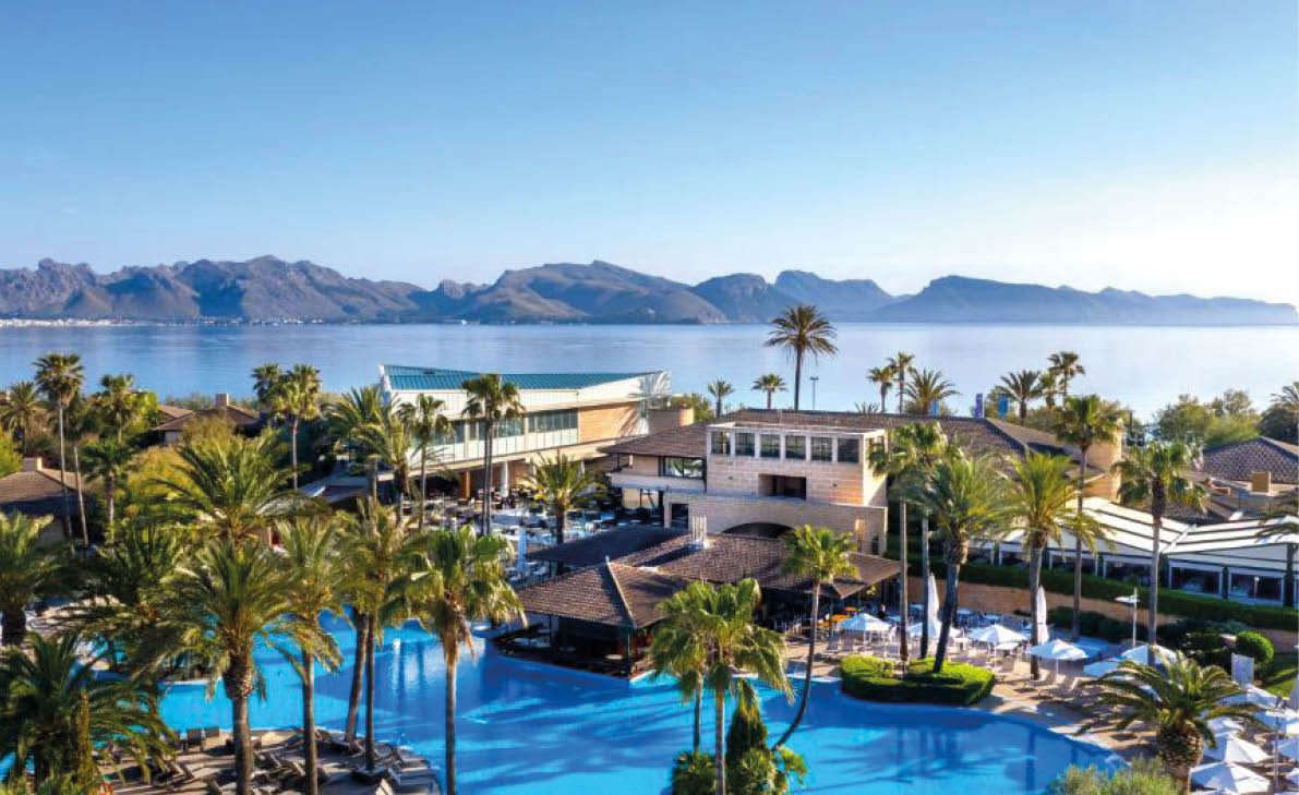 A luxurious resort with a large blue swimming pool surrounded by palm trees and buildings, set against a backdrop of mountains and a calm sea under a clear sky. Beschreibung von KI generiert