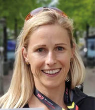 A person with long blonde hair is smiling outdoors wearing a black jacket and sunglasses on their head with a lanyard around their neck. Beschreibung von KI generiert