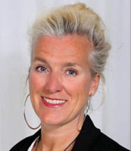 A person with light-colored hair styled up is smiling and wearing large hoop earrings and a black top against a plain light background. Beschreibung von KI generiert