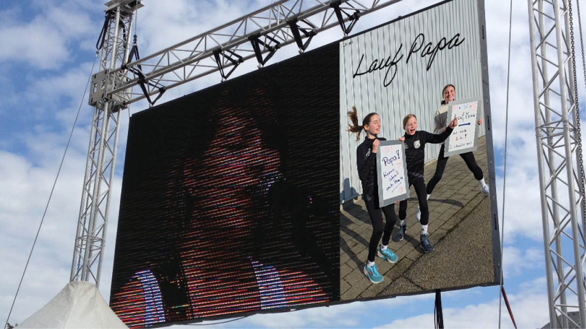 A large outdoor screen displays a close-up of a person singing while a smaller inset shows three people holding signs and energetically posing against a corrugated metal wall under a partly cloudy sky. Beschreibung von KI generiert