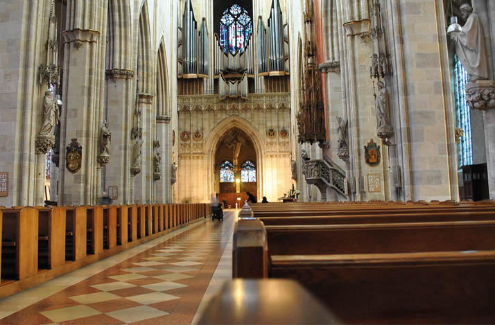 A large, ornate cathedral interior with wooden pews, tall stone columns, stained glass windows, and an organ visible at the far end. Beschreibung von KI generiert