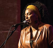 A person with curly hair wearing a yellow headband and a brown leather jacket is singing into a microphone on stage. Beschreibung von KI generiert