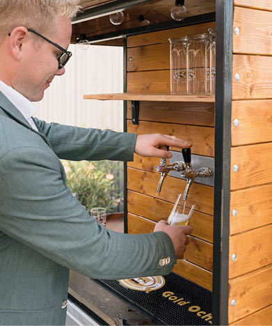A person wearing glasses and a suit jacket is pouring beer from a tap into a glass at a wooden bar counter with shelves holding empty glasses. Beschreibung von KI generiert