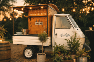 A small white vintage truck converted into a rustic outdoor bar with wooden panels, taps, hanging glasses, string lights, and surrounded by potted plants and candles at sunset. Beschreibung von KI generiert