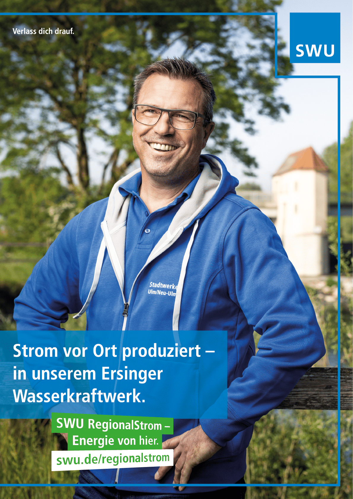 A smiling person wearing glasses and a blue jacket stands outdoors near a wooden fence with trees and a building in the background, accompanied by text promoting local electricity production from a hydropower plant. Beschreibung von KI generiert