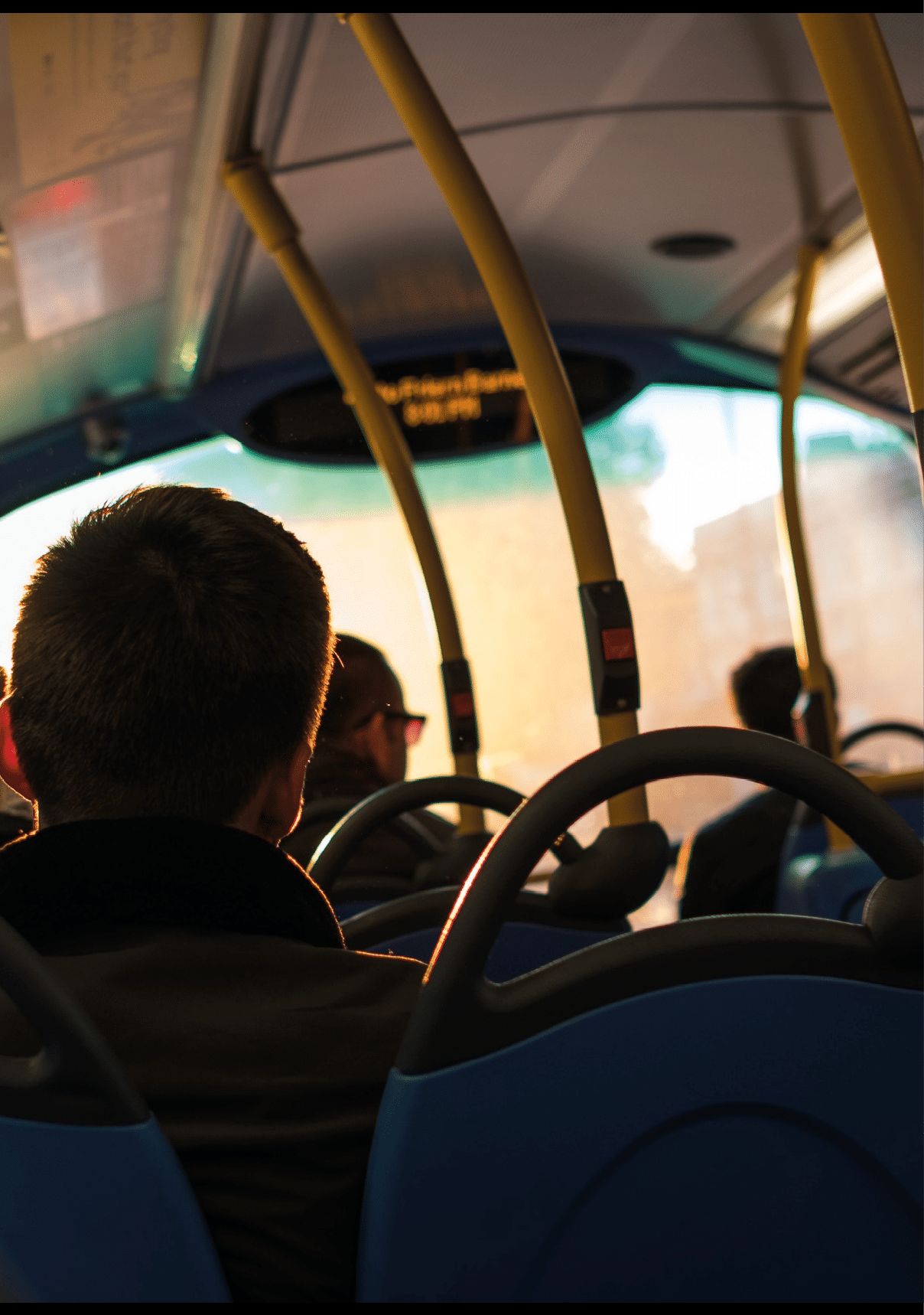 People are seated inside a bus with sunlight streaming through the windows, highlighting the backs of their heads and the blue seats. Beschreibung von KI generiert