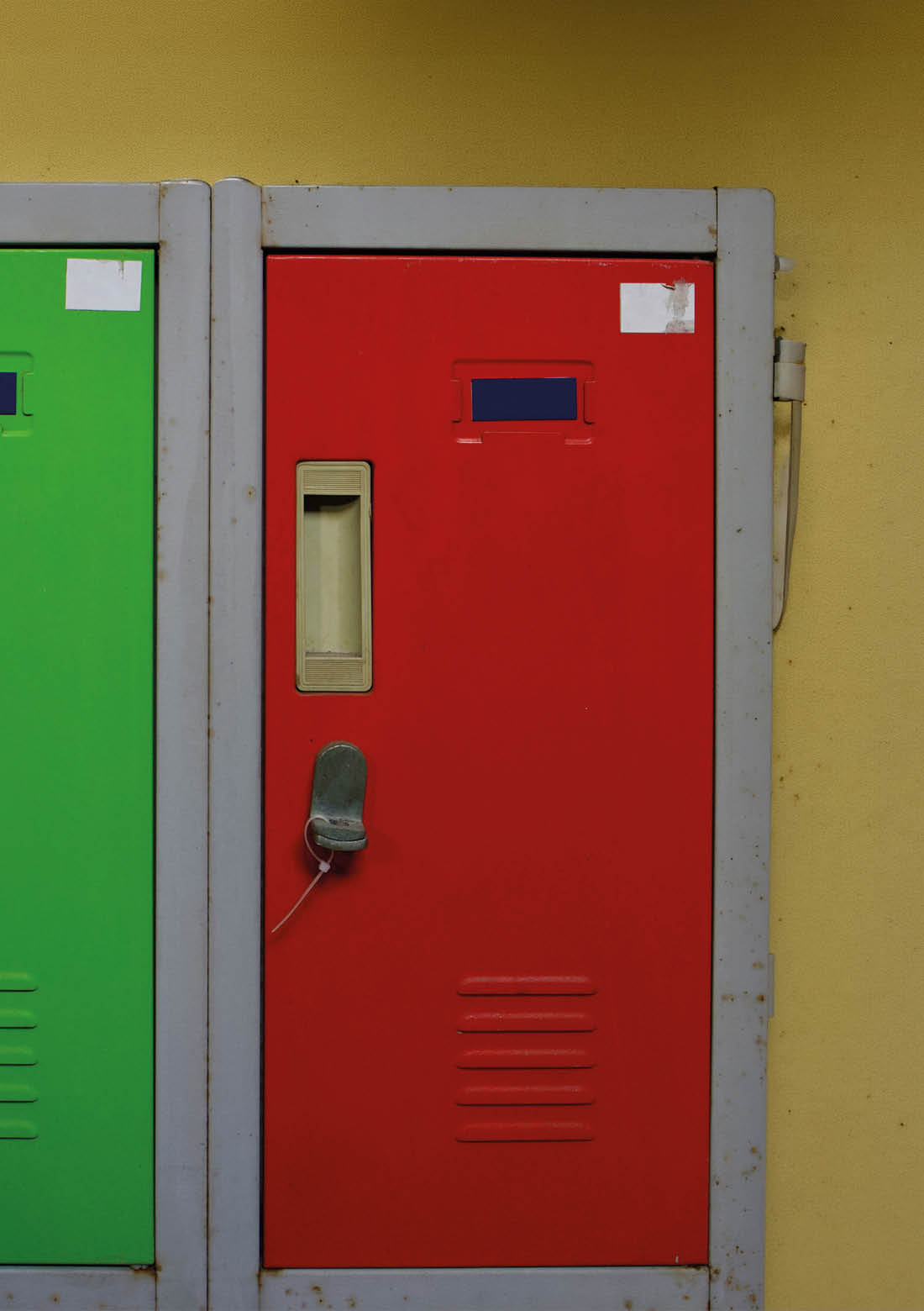 A red metal locker with a beige handle and a small lock hanging from it is positioned next to a green locker against a yellow wall. Beschreibung von KI generiert