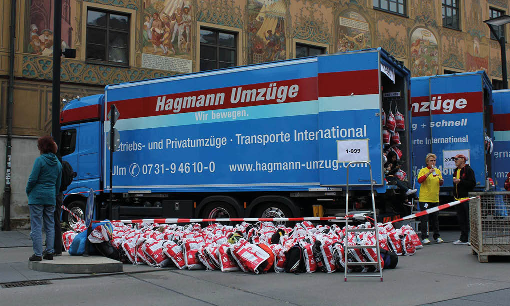 A group of people stands near a blue and red moving truck with many red and white bags arranged on the ground in front of it, set against a building with detailed wall murals. Beschreibung von KI generiert