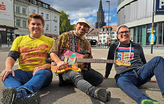 Three adults sitting on the ground in a city square with one holding a colorful electric guitar, all smiling and dressed casually. Beschreibung von KI generiert