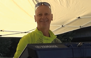 A person wearing a bright green jacket and sunglasses on their head is smiling while standing behind a Yamaha keyboard under a canopy. Beschreibung von KI generiert