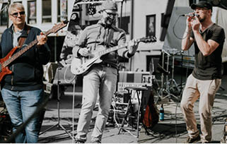 Three adults are performing music outdoors with one playing a bass guitar, another playing an electric guitar, and the third singing into a microphone. Beschreibung von KI generiert