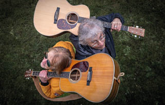 Two adults with gray and light brown hair are sitting back-to-back on grass, each holding an acoustic guitar, one wearing a yellow jacket and sunglasses and the other wearing a dark jacket and glasses. Beschreibung von KI generiert