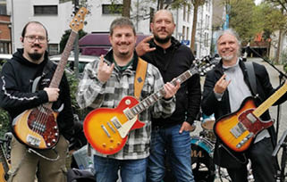 Four adults stand outdoors holding electric guitars and making rock hand gestures while smiling. Beschreibung von KI generiert