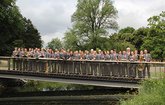 A large group of people dressed in matching suits stand closely together on a bridge over a body of water surrounded by trees and greenery. Beschreibung von KI generiert