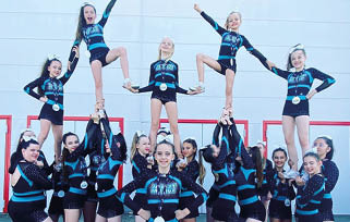 A group of people in matching blue and black cheerleading uniforms with bows and medals are posing in a cheerleading stunt formation against a light-colored wall. Beschreibung von KI generiert