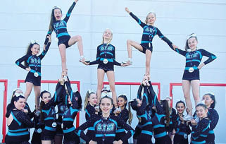 A group of people in matching blue and black cheerleading uniforms are performing a coordinated stunt with some standing on others' shoulders and holding hands against a plain wall background. Beschreibung von KI generiert