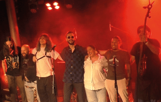 A group of seven adults stand closely together on a stage with red lighting, smiling and posing with musical instruments and microphones. Beschreibung von KI generiert