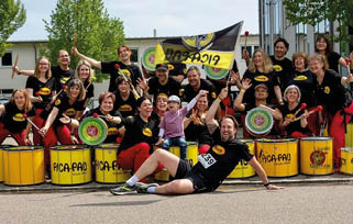 A group of adults wearing matching black shirts and red pants are posing happily with yellow drums and drumsticks outdoors, with one person lying in front and another holding a flag. Beschreibung von KI generiert