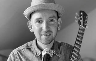 A person wearing a hat and denim shirt is smiling while holding a guitar and standing behind a microphone. Beschreibung von KI generiert